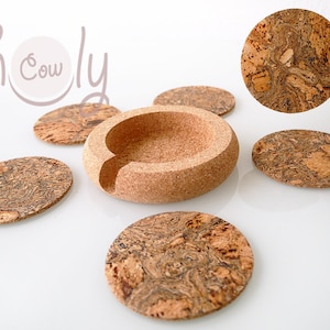 Puede incluir: Posavasos y un soporte de corcho natural. Los posavasos son redondos y ovalados, con un patrón único. El soporte tiene una forma redondeada con una abertura curva. La imagen incluye el texto "h Cowly".