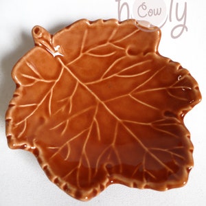 K&ouml;nnte beinhalten: Eine braune, blattf&ouml;rmige Keramikschale mit gl&auml;nzendem Finish. Die Schale hat detaillierte Adernmuster, die einem Herbstblatt &auml;hneln. Ideal zum Servieren von Snacks oder als dekoratives Element. Das Wort "holy cow" ist kursiv geschrieben.