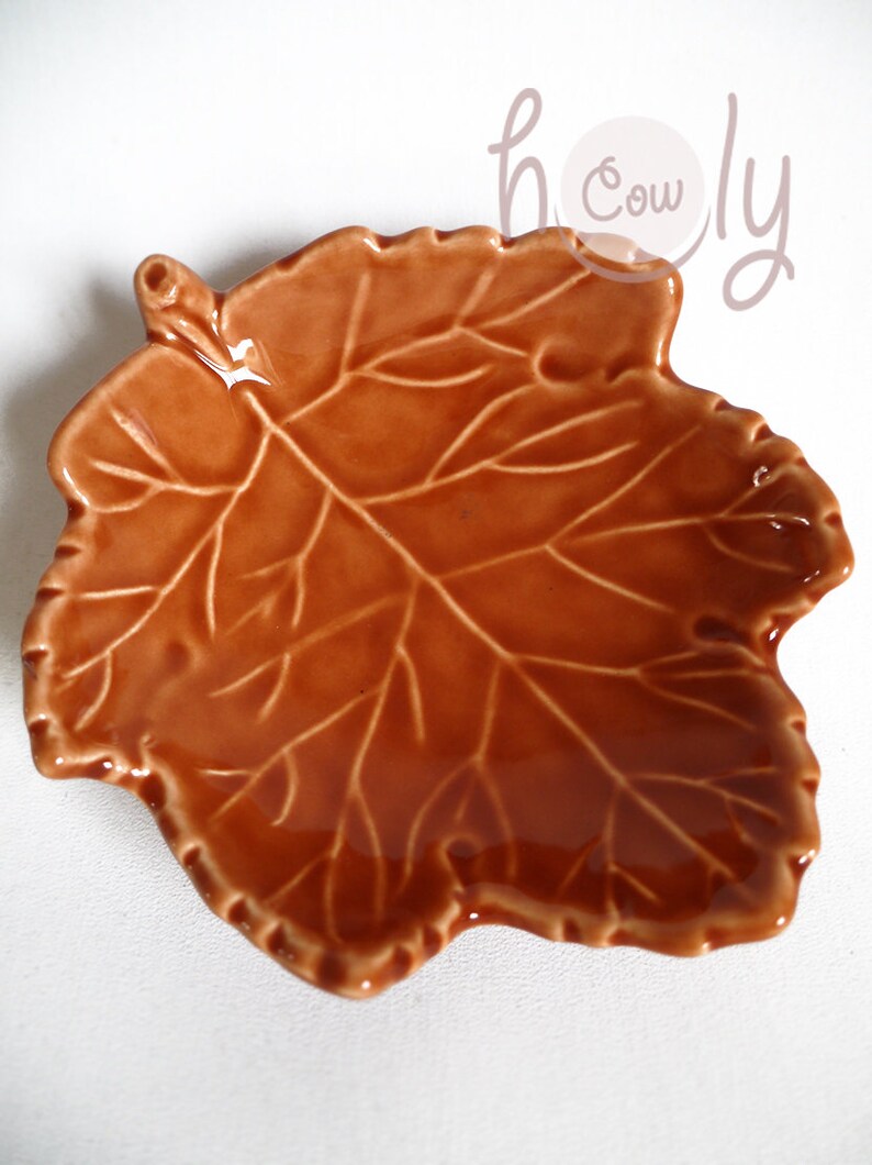 K&ouml;nnte beinhalten: Eine braune, blattf&ouml;rmige Keramikschale mit gl&auml;nzendem Finish. Die Schale hat detaillierte Adernmuster, die einem Herbstblatt &auml;hneln. Ideal zum Servieren von Snacks oder als dekoratives Element. Das Wort "holy cow" ist kursiv geschrieben.