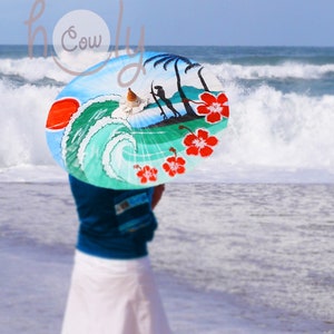 Op de afbeelding: Een vrouw op een strand die een kleurrijke parasol vasthoudt met een golf en palmbomen erop geschilderd. De parasol is geschilderd met rode hibiscusbloemen.
