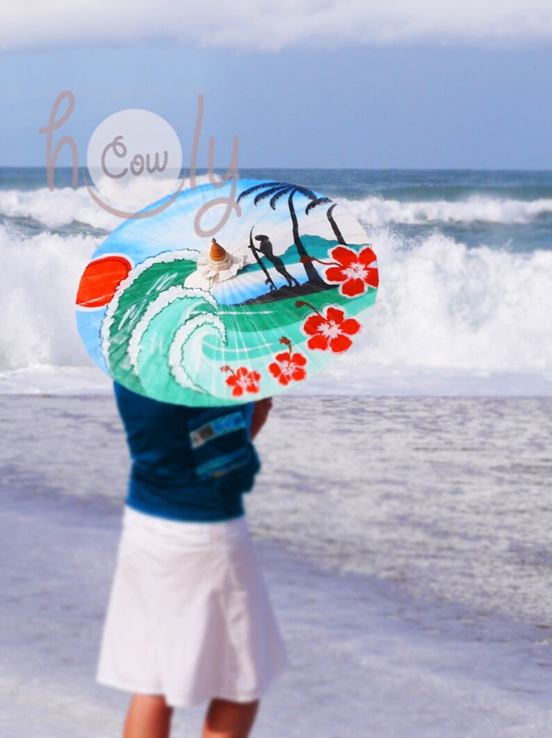 Op de afbeelding: Een vrouw op een strand die een kleurrijke parasol vasthoudt met een golf en palmbomen erop geschilderd. De parasol is geschilderd met rode hibiscusbloemen.