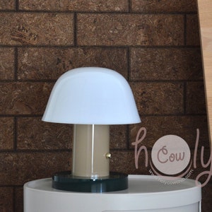 Könnte beinhalten: Eine weiße pilzförmige Lampe mit einem beigefarbenen Sockel und einem dunkelgrünen Sockel. Die Lampe steht auf einem weißen Nachttisch.