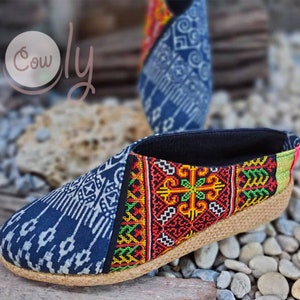 K&ouml;nnte beinhalten: Ein Paar blaue und schwarze Slipper mit einem Obermaterial aus gewebtem Stoff und einer Jutesohle. Die Schuhe haben ein farbenfrohes Stickmuster an der Seite.