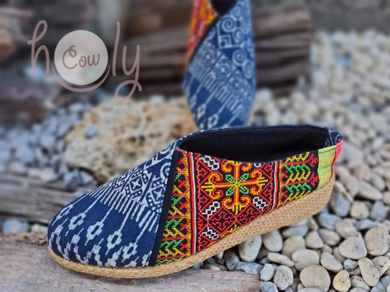 K&ouml;nnte beinhalten: Ein Paar blaue und schwarze Slipper mit einem Obermaterial aus gewebtem Stoff und einer Jutesohle. Die Schuhe haben ein farbenfrohes Stickmuster an der Seite.