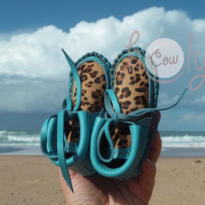 May include: A pair of turquoise leather moccasins with leopard print fabric lining. The moccasins are tied together with the laces.