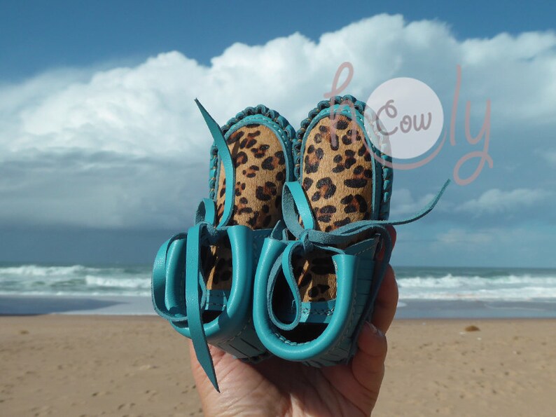 May include: A pair of turquoise leather moccasins with leopard print fabric lining. The moccasins are tied together with the laces.