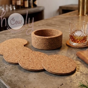 Pode incluir: Uma coleção de bases de cortiça e uma tigela de cortiça em uma superfície de concreto. Um copo de líquido âmbar com um grande cubo de gelo está em uma base. Um jigger de cobre e um caderno de couro também estão presentes.