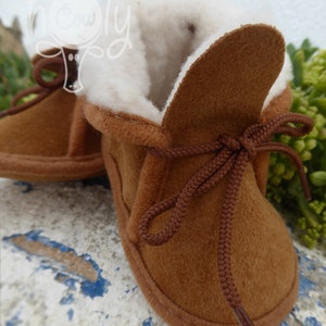 May include: Brown suede baby booties with a soft, white interior lining. The booties have a lace-up design with brown laces and a rounded toe. These cozy baby shoes are designed for comfort and warmth.