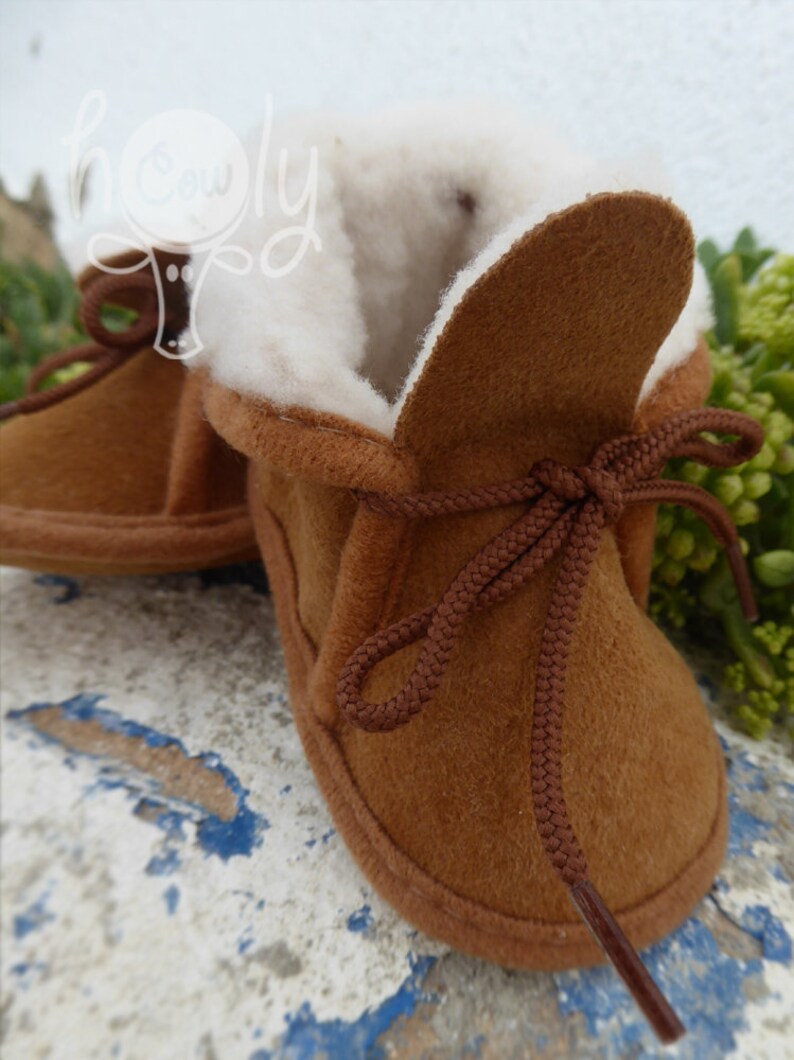 May include: Brown suede baby booties with a soft, white interior lining. The booties have a lace-up design with brown laces and a rounded toe. These cozy baby shoes are designed for comfort and warmth.