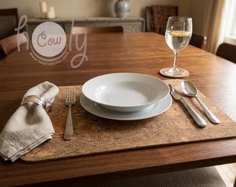 May include: A dining table setting with a white bowl and plate, silverware, a linen napkin with a silver ring, and a glass of water with a lemon slice. The setting is on a cork placemat.