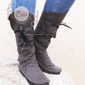 May include: A pair of gray leather boots with a flat sole and a knee-high design. The boots feature wraparound straps, white stitching, and a folded-over top. The boots are shown against a light background.