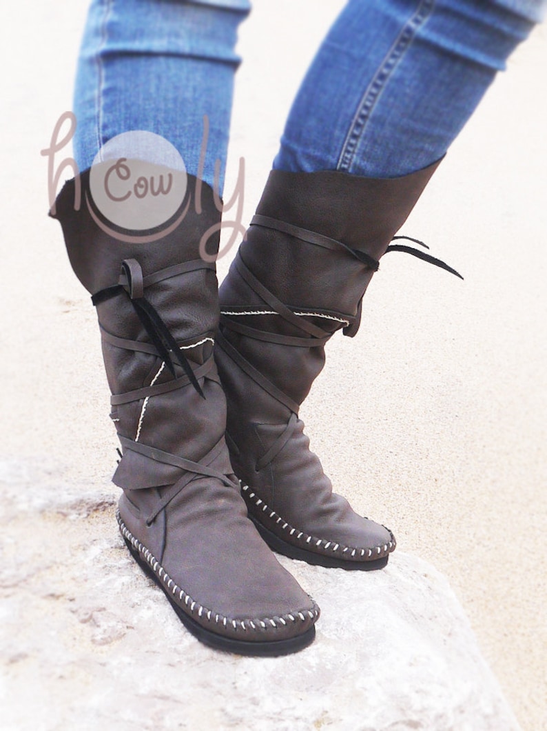 May include: A pair of gray leather boots with a flat sole and a knee-high design. The boots feature wraparound straps, white stitching, and a folded-over top. The boots are shown against a light background.