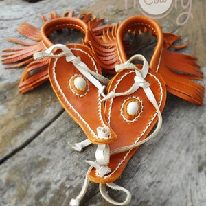 May include: Two orange leather wrist cuffs with white stitching and fringe. The cuffs have a white leather strap and a white bead in the center. The cuffs are on a wooden surface. The text "holy cow" is visible in the background.