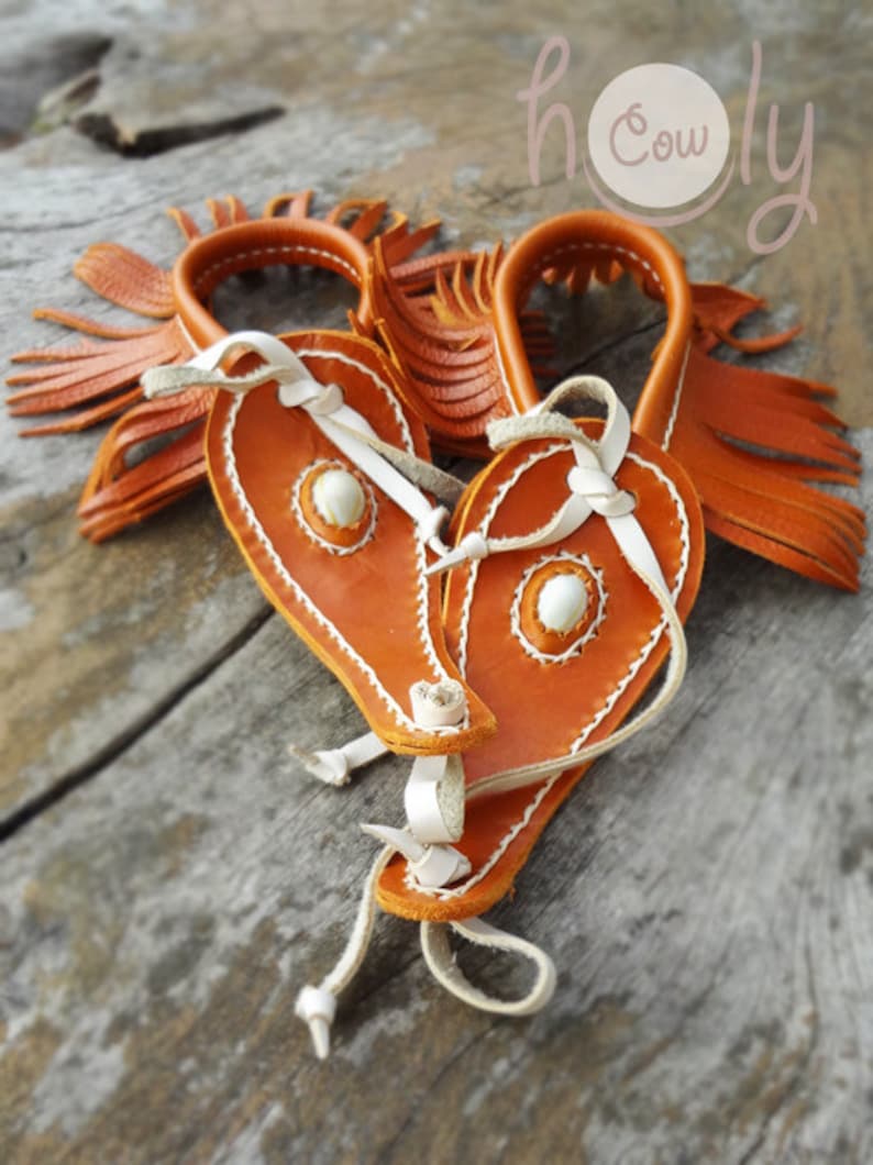 May include: Two orange leather wrist cuffs with white stitching and fringe. The cuffs have a white leather strap and a white bead in the center. The cuffs are on a wooden surface. The text "holy cow" is visible in the background.