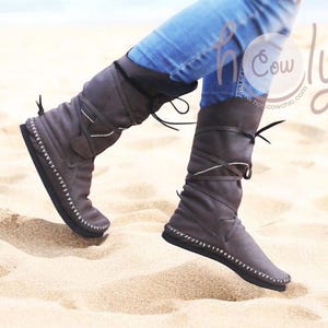 May include: Gray knee-high boots with a flat sole and white stitching. The boots have a lace-up design with a strap detail and are worn with blue jeans. The boots are made of a soft material.
