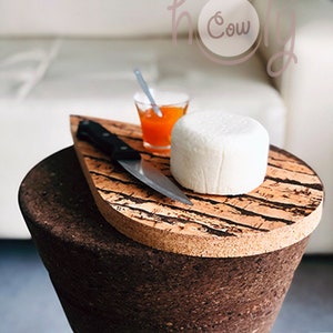 Może przedstawiać: Okrągły biały ser na korkowej desce do serwowania z małym nożem i szklanką pomarańczowej marmolady. Deska ma kształt łzy i wzór słojów drewna. Deska stoi na korkowym stołku.