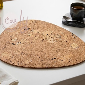 Op de afbeelding: Een grote, ovale kurken placemat met een natuurlijk, getextureerd oppervlak. De placemat is lichtbruin met donkerbruine en zwarte markeringen. Een donkere koffiemok staat op een kleine, zwarte onderzetter met een lepel.