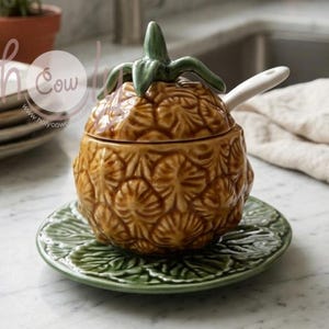 Peut inclure: Un plat de service en céramique en forme d'ananas avec un couvercle et une petite cuillère. Le plat est marron avec une surface texturée, posé sur une assiette verte en forme de feuille. La couronne de l'ananas est verte.