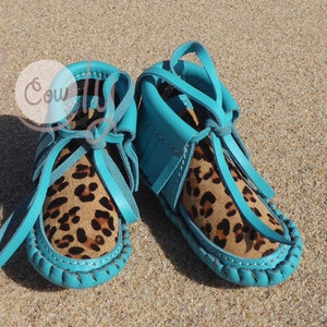 May include: A pair of turquoise blue baby moccasins with leopard print leather uppers and fringe detail.