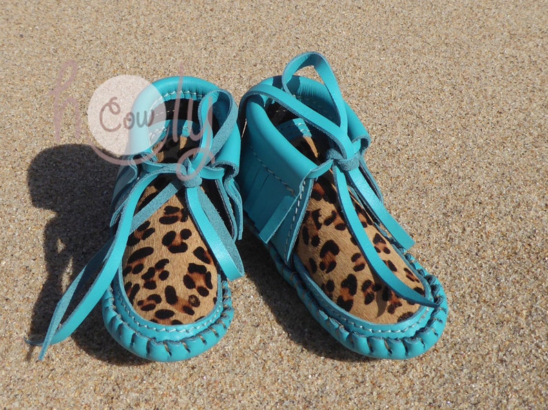May include: A pair of turquoise blue baby moccasins with leopard print leather uppers and fringe detail.