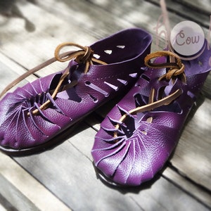 May include: A pair of purple leather sandals with brown leather laces. The sandals have a unique design with a folded leather detail at the toe.