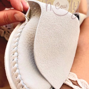 May include: Close-up of a white leather shoe being sewn with a needle and thread. The shoe has a decorative stitch around the edge and a flap of leather covering the top.