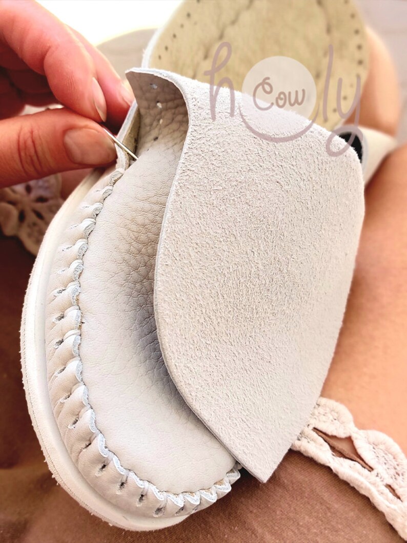 May include: Close-up of a white leather shoe being sewn with a needle and thread. The shoe has a decorative stitch around the edge and a flap of leather covering the top.
