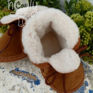 May include: A pair of brown baby booties with a soft, white interior and trim. The booties have brown laces and are made of a suede-like material. The image is set against a background of green foliage and a distressed, blue and white surface.