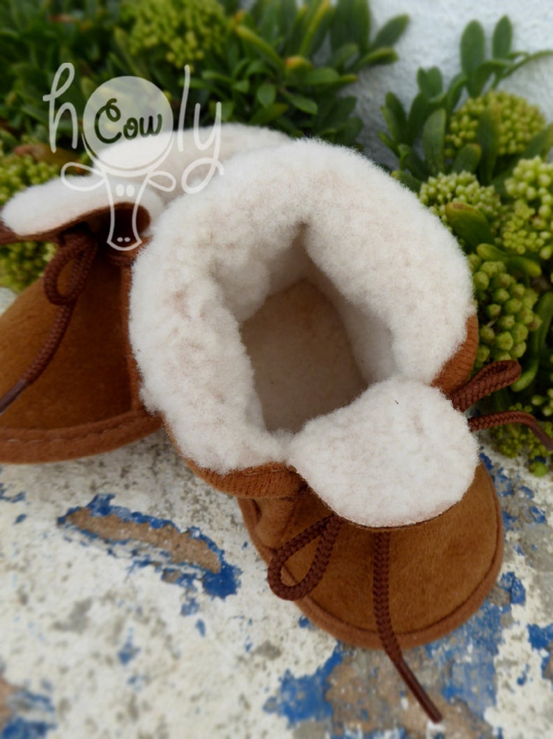 May include: A pair of brown baby booties with a soft, white interior and trim. The booties have brown laces and are made of a suede-like material. The image is set against a background of green foliage and a distressed, blue and white surface.