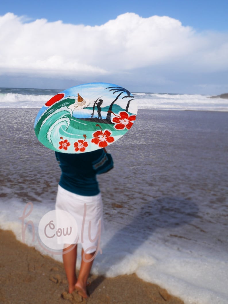 Op de afbeelding: Een vrouw staat op een strand en houdt een kleurrijke paraplu vast met een geschilderd tafereel van een surfer die op een golf rijdt, palmbomen en hibiscusbloemen.