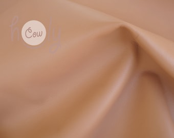 May include: A close-up of a light brown leather fabric with a subtle texture. The fabric is folded and draped, showcasing its soft and supple quality.