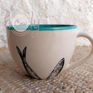May include: Cream-colored ceramic mug with a teal rim and a white handle. The mug features a black and white illustration of two sardines. The text "holy cow chic" is visible on the side of the mug.