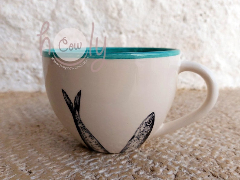 May include: Cream-colored ceramic mug with a teal rim and a white handle. The mug features a black and white illustration of two sardines. The text "holy cow chic" is visible on the side of the mug.