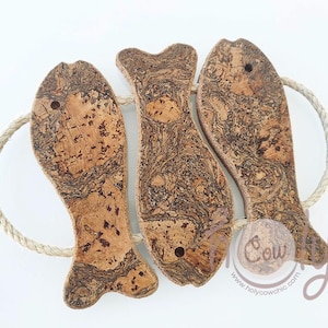 May include: Three cork fish-shaped hot pads with a rope handle. The hot pads are brown with a marbled pattern. The text "Holy Cow Chic" is printed on the bottom of the hot pads.