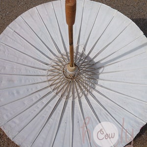 May include: A white paper parasol with a wooden handle and frame. The parasol is open, showing its radiating ribs and central design. The handle is light brown, and the paper has a slightly textured appearance.
