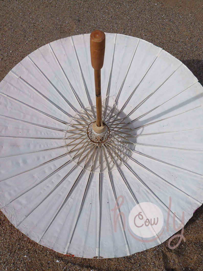 May include: A white paper parasol with a wooden handle and frame. The parasol is open, showing its radiating ribs and central design. The handle is light brown, and the paper has a slightly textured appearance.