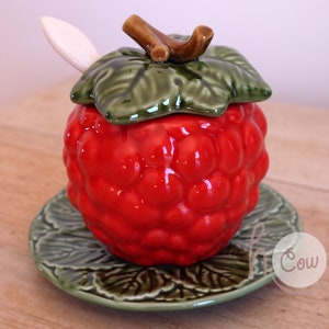 Puede incluir: Un azucarero de cerámica rojo en forma de frambuesa con una tapa de hoja verde y una pequeña cuchara. El bol se asienta sobre un platillo de hoja verde a juego.