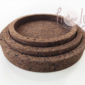 May include: Three round cork trays of varying sizes are stacked on top of each other. The trays are a natural brown color with a textured surface. The trays have a raised edge, creating a shallow bowl shape. The background is white.