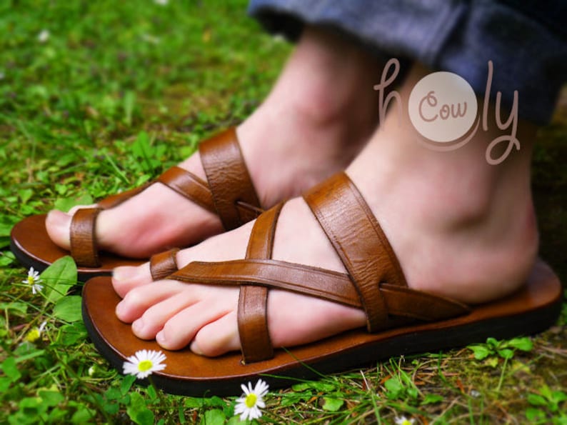 May include: Brown leather sandals with multiple straps crossing over the foot. The sandals are worn by a person with the text "Holy Cow" tattooed on their ankle.