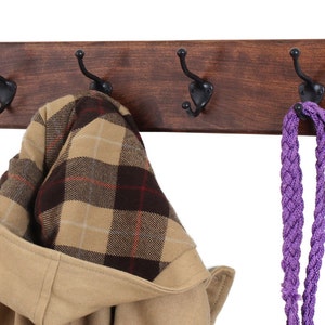 May include: A wooden coat rack with three black metal hooks. A brown jacket with a plaid lining is hanging on one hook, and a purple braided rope is hanging on another hook.