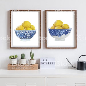 Könnte beinhalten: Zwei gerahmte Drucke von Aquarellmalereien von Zitronen in blauen und weißen Schalen. Die Drucke hängen an einer weißen Wand über einer weißen Kommode mit einem Holzregal und drei kleinen Topfpflanzen. Die Kommode hat auch eine schwarze Gießkanne und ein weißes Schild mit der Aufschrift "Sei positiv".