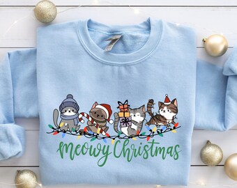 Peut inclure: Un sweat-shirt bleu clair avec un motif de Noël représentant quatre chats portant des bonnets de Père Noël et tenant des cadeaux. Le texte "Meowy Christmas" est imprimé en vert sous les chats.