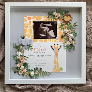 May include: A white frame with a baby shower invitation inside. The invitation is for a baby shower honoring Maura Negri. The invitation has a giraffe, an elephant, and a monkey on it. The text on the invitation reads "A Little Wild One is on the Way".
