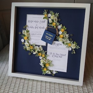 Könnte beinhalten: Ein weißer Rahmen mit einem dunkelblauen Hintergrund und einer Papierblumen-Arrangement in Gelb und Weiß. Der Rahmen enthält eine Hochzeitseinladung für Kristin Marie Sulaff und Nicholas Kent Meader. Die Einladung enthält das Datum der Hochzeit, den 06.02.2018, und den Ort der Rezeption, das St. Louis Carousel im Forest Park.