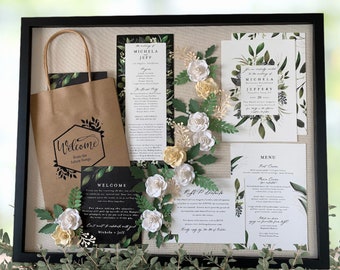May include: A framed wedding invitation suite featuring a kraft paper gift bag with "Welcome" text, invitation cards, and a menu. The suite includes white and yellow paper flowers and greenery. The invitation suite is displayed in a black frame.