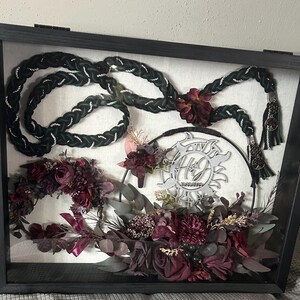 May include: A rectangular black shadow box frame containing a braided dark green and white cord, a circular metal emblem with the letters HJ and the date 10-29-2024, and an arrangement of deep burgundy and dark purple artificial flowers and foliage.