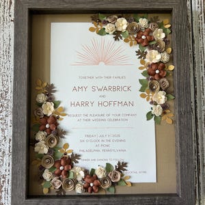 May include: A framed wedding invitation featuring the names Amy Swarbrick and Harry Hoffman. The invitation is surrounded by paper flowers in shades of brown, cream, and green. The frame is a weathered gray color. The invitation is for a wedding on Friday, July 11, 2025.
