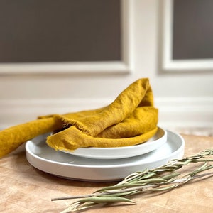 May include: A mustard yellow linen napkin folded and placed on top of two white plates. The plates are on a wooden surface with a sprig of dried greenery.
