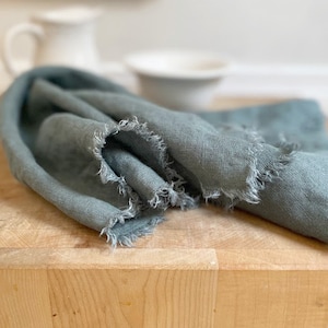 Puede incluir: Una toalla de cocina de lino azul suave con bordes deshilachados. La toalla est&aacute; doblada y colocada sobre una tabla de cortar de madera.