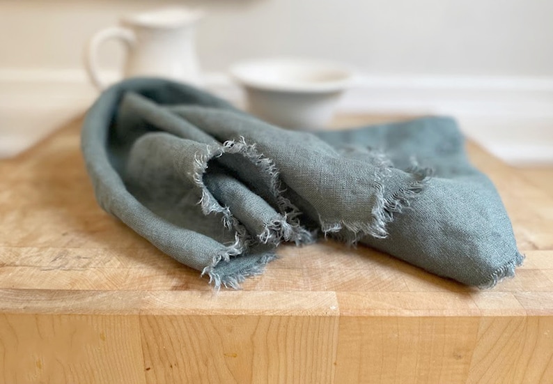 Puede incluir: Una toalla de cocina de lino azul suave con bordes deshilachados. La toalla est&aacute; doblada y colocada sobre una tabla de cortar de madera.